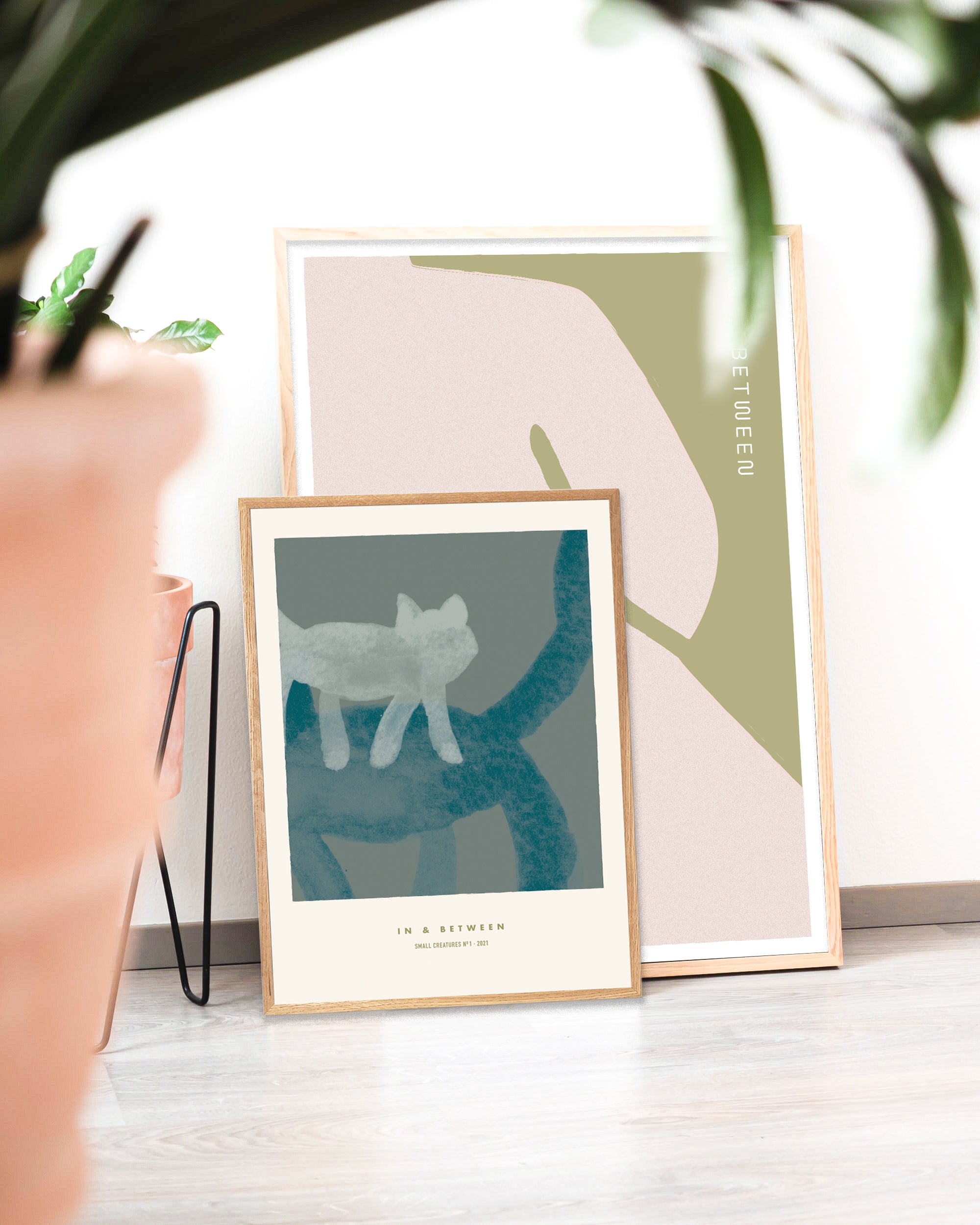 Two framed art prints leaning agains a wall. Largest print is an abstraction of a sitting female form in light red/olive green and smaller print in front shows a small light grey cat perched on back of larger green cat on grey background.