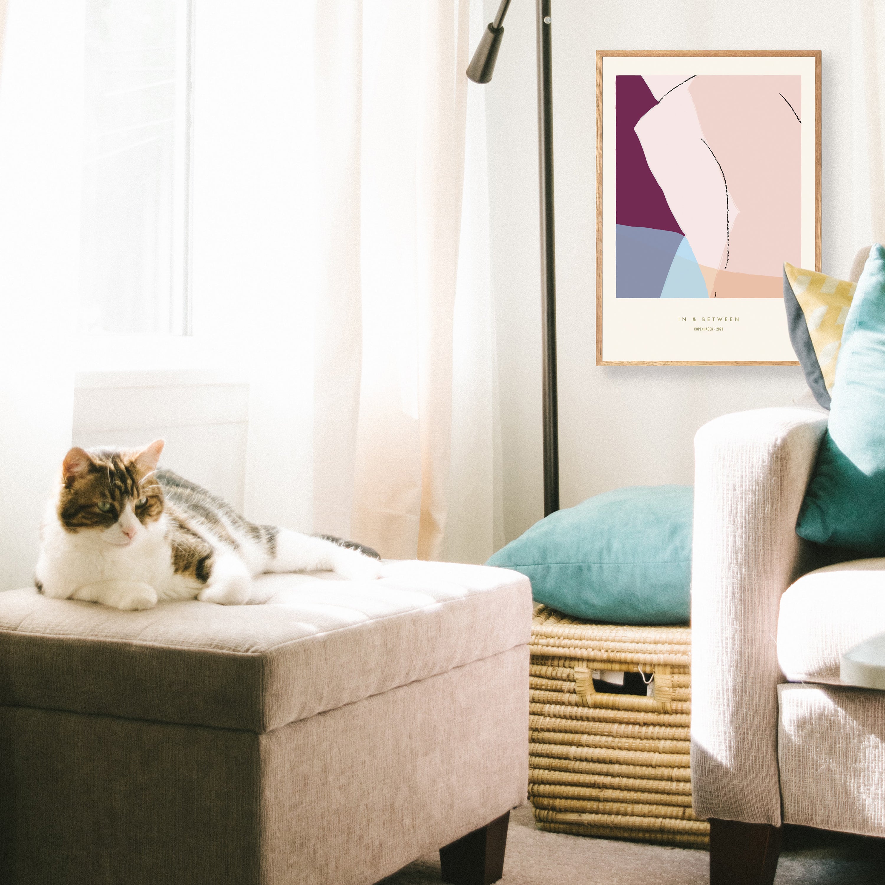 Wooden framed art print of lightly abstracted human turning in overlapping transparent colours berry, pale pink, grey/blue hung on wall in living room with a cat on a pouffe, a basket and a sofa.